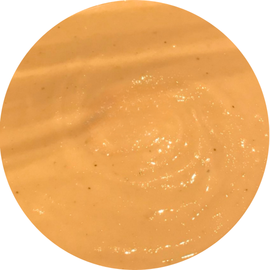 A circular close-up swatch of a smooth, glossy orange-yellow creamy texture of cashew butter with subtle swirls and tiny dark flecks, set against a plain white background.