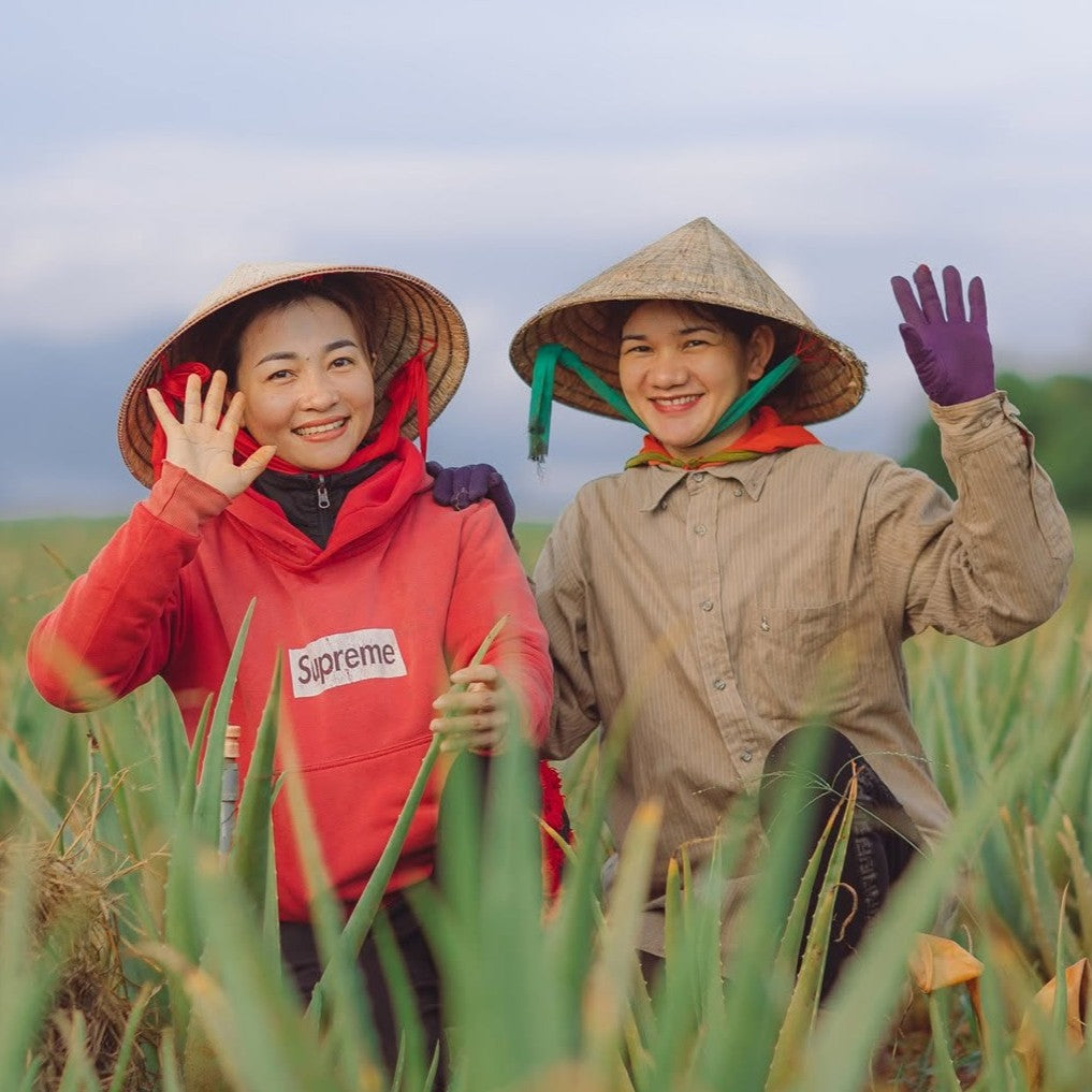 Two people smiling and waving in a field while wearing conical hats and work clothes. One person wears a red hoodie with a visible Supreme logo, and the other wears a beige shirt with purple gloves.
